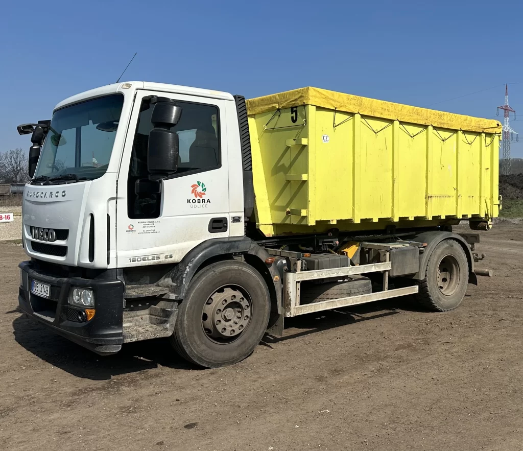 Nákladní vůz Iveco Eurocargo bílé barvy s připojeným žlutým kontejnerem. Na dveřích vozu je logo a název 'Kobra Udlice'. Kontejner je velký, obdélníkový, a má označení číslem 5.