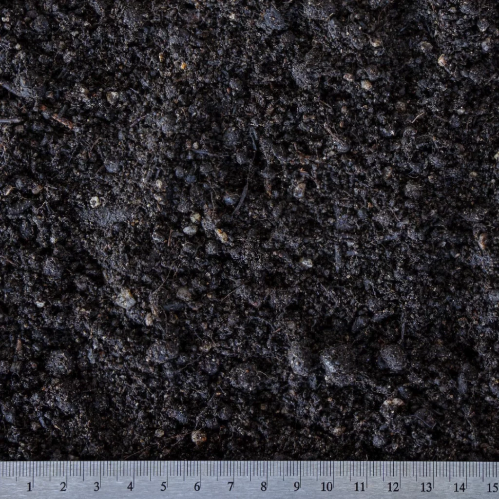 Detailní pohled na zahradnický substrát frakce 0–20 mm s viditelnými organickými částicemi a drobnými kamínky, doplněný měřítkem pro určení velikosti zrn.